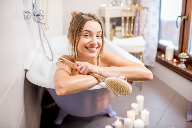 Porträt einer schönen Frau, die mit Bürste in der Retro-Badewanne im Vintage-Badezimmer mit Kerzen liegt