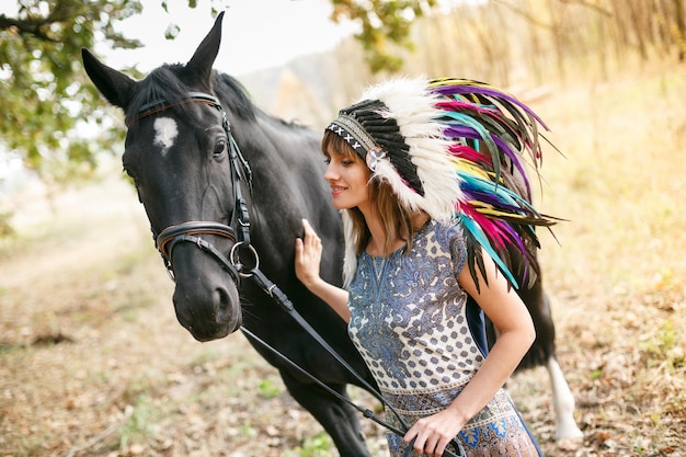 Porträt einer schönen Frau in einem Kleid und indianischen Federn mit einem schwarzen Pferd im Freien Tagesaufnahme