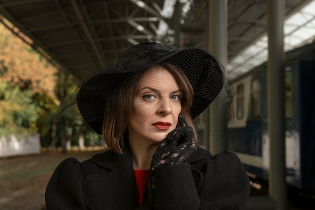 Porträt einer schönen Frau in Retro-Kleidung und schwarzem Hut mit breiter Krempe, die auf dem Bahnsteig des Bahnhofs steht