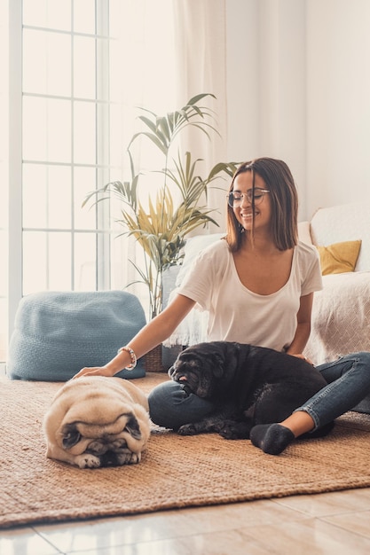 Porträt einer schönen Frau mit Brille, die Spaß mit ihren Haustieren hat, Hunde, die auf dem Boden im Wohnzimmer ihres Hauses sitzen. Fröhliche Frau verbringt Freizeit mit ihrem Hund mit zwei süßen Haustieren zu Hause