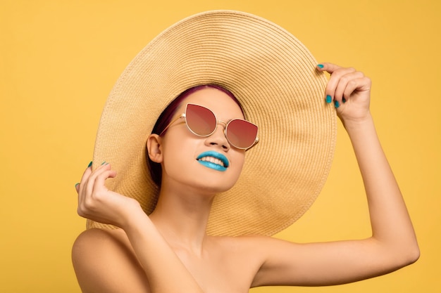 Porträt einer schönen Frau mit hellem Make-up-Hut und Sonnenbrille auf gelbem Studiohintergrund