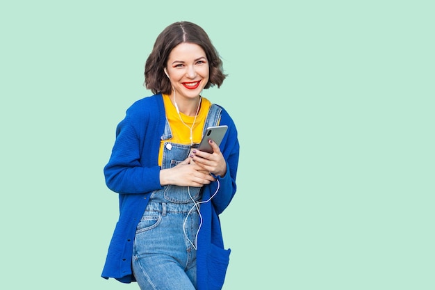 Porträt einer schönen, fröhlichen jungen erwachsenen Frau in Hipster-Kleidung in Denim-Overalls, die steht, Telefon hält und Lieblingsmusik mit Hörern hört. Studioaufnahme, grüner Hintergrund, Blick in die Kamera