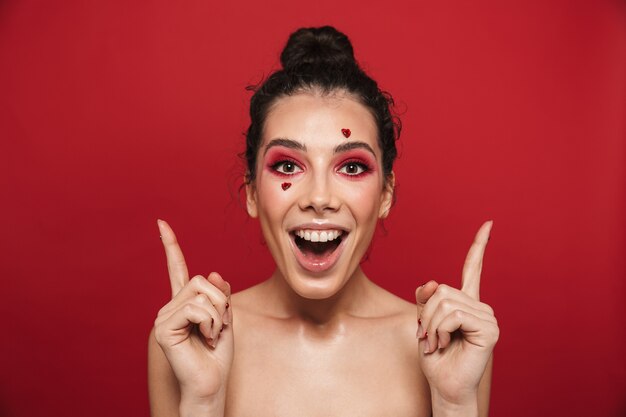 Porträt einer schönen glücklichen fröhlichen jungen Frau mit rotem hellem Make-up einzeln auf roter Wand, die mit Herzen auf dem Gesicht aufwirft, das auf Exemplar zeigt.