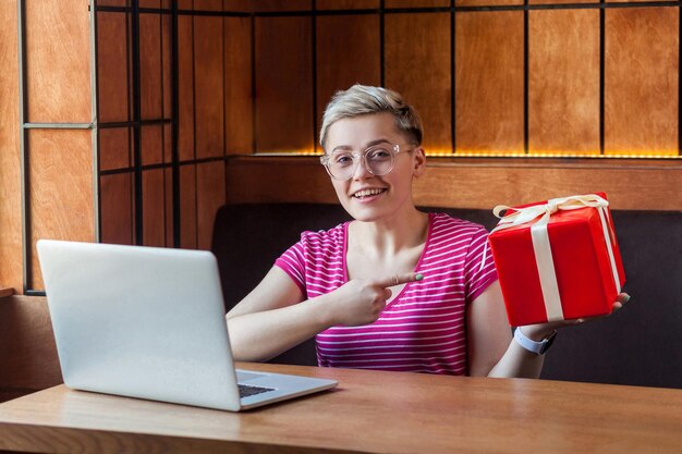 Porträt einer schönen glücklichen jungen Frau mit kurzen Haaren in rosafarbenem T-Shirt sitzt im Café und zeigt mit dem Finger auf eine rote Geschenkbox. Einfach online zu bestellen