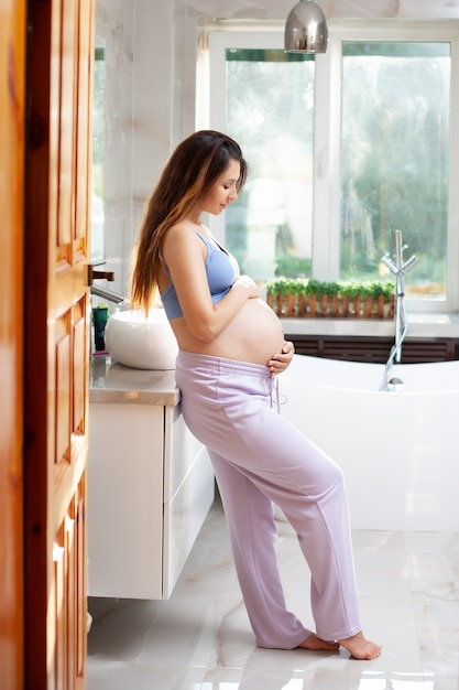 Porträt einer schönen glücklichen schwangeren Brünette in einem hellen Badezimmer. Morgen Sonnenschein. Lächeln, Glück. Vertikal. Foto in hoher Qualität
