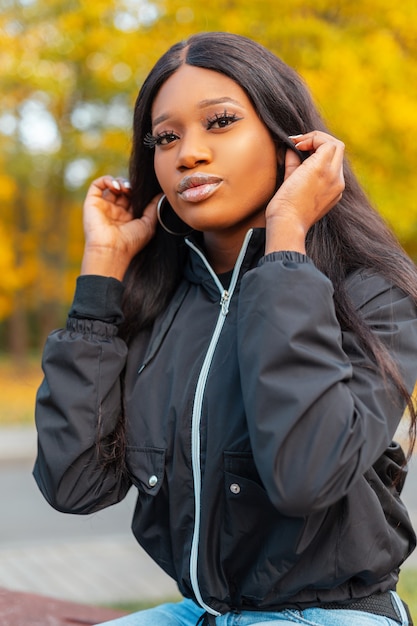 Porträt einer schönen jungen afroamerikanischen Frau mit einer schwarzen modischen Jacke in der Natur auf einem Hintergrund von gelbem Herbstlaub