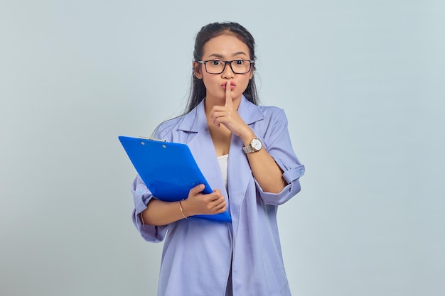 Porträt einer schönen jungen asiatischen Frau, die einen Dokumentenordner hält und eine stille Geste macht, die auf violettem Hintergrund isoliert ist