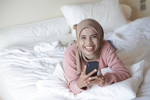 Porträt einer schönen jungen asiatischen muslimischen Frau mit Hijab auf dem Bett, die ihr Mobiltelefon in der Hand hält