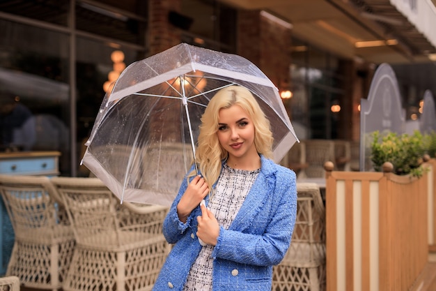 Porträt einer schönen jungen blonden Frau, die einen durchsichtigen Regenschirm im Regen auf einer Stadtstraße hält