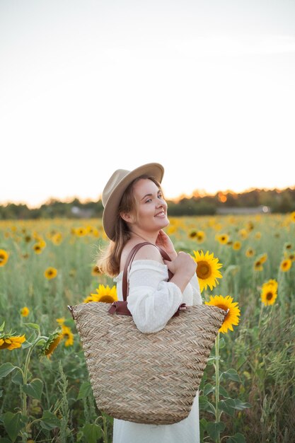 Porträt einer schönen jungen Frau, 33 Jahre alt, mit Hut im Sonnenblumenfeld bei Sonnenuntergang Glückliches Modell im weißen Kleid am Sommerabend in der Natur Warm