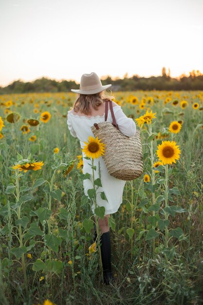 Porträt einer schönen jungen Frau, 33 Jahre alt, mit Hut im Sonnenblumenfeld bei Sonnenuntergang Glückliches Modell im weißen Kleid am Sommerabend in der Natur Warm