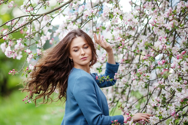 Porträt einer schönen jungen Frau der Brünette in der blauen Jacke im blühenden Apfelbaumgarten im Frühling. Natur genießen. Gesundes Mädchen im Freien. Frühlingskonzept. Schönes Mädchen im Obstgarten