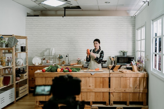 Porträt einer schönen jungen Frau, die Gemüse an der Küchentheke schneidet. Familienhausfrau, die ein Video zum Kochen eines Blogs aufzeichnet. Vlogger macht Salat Schneiden frischer Karotten im Gespräch mit Kamera auf Stativ