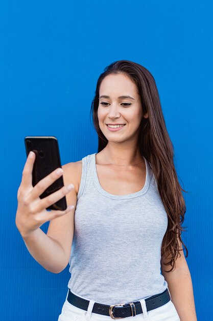 Porträt einer schönen jungen Frau, die lächelt und ein Smartphone mit einer blauen Wand im Hintergrund verwendet