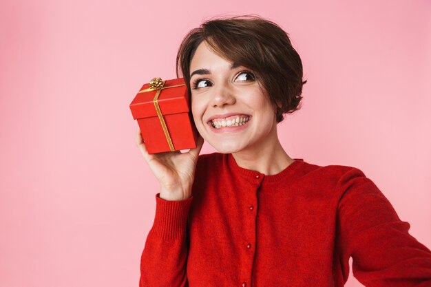 Porträt einer schönen jungen Frau, die rote Kleidung trägt, die lokal über rosa Hintergrund steht und Geschenkbox zeigt