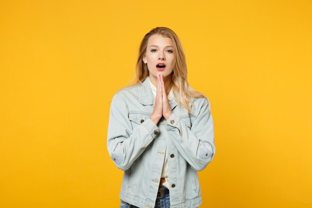 Porträt einer schönen jungen Frau in Denim-Freizeitkleidung, die in die Kamera blickt, die Hände gefaltet hält und isoliert auf gelb-orangefarbenem Hintergrund im Studio betet. People-Lifestyle-Konzept. Mock-up-Kopienbereich.