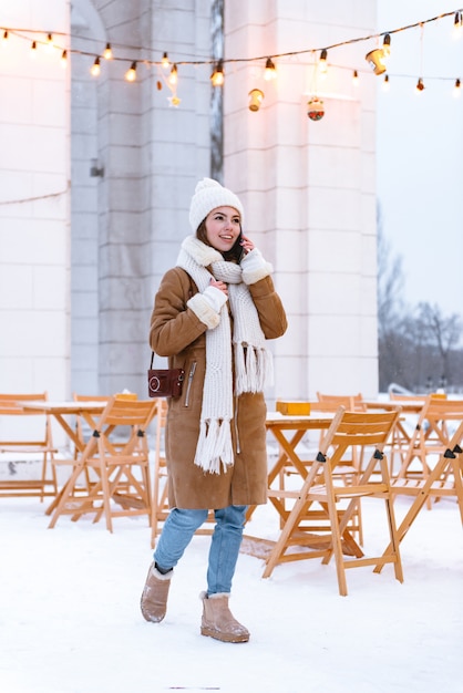 Porträt einer schönen jungen Frau in der Mütze und im Schal, die draußen im Winterschnee sprechend durch Handy sprechen.