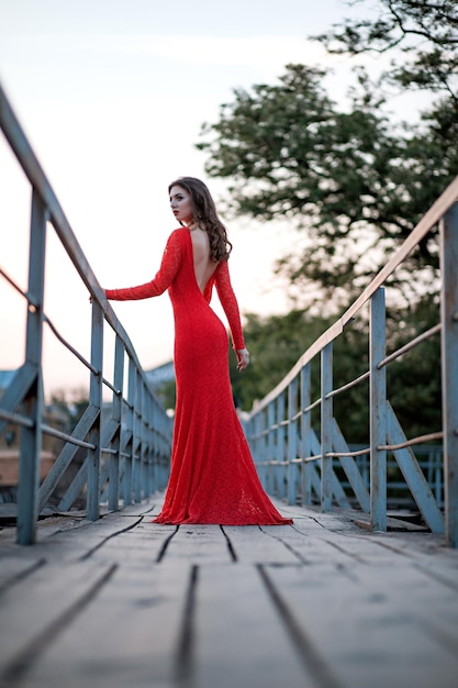 Porträt einer schönen jungen Frau in einem langen roten Kleid steht auf der Brücke