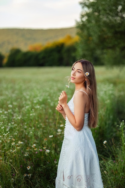 Porträt einer schönen jungen Frau in einem weißen Sommerkleid mit einer Kamille in ihren Händen Sommerspaziergang in den Bergen