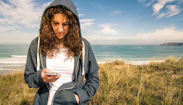 Porträt einer schönen jungen Frau mit blauem Hoodie und Sportbekleidung, die ihr Smartphone über einem bewölkten Hintergrund des blauen Himmels schaut