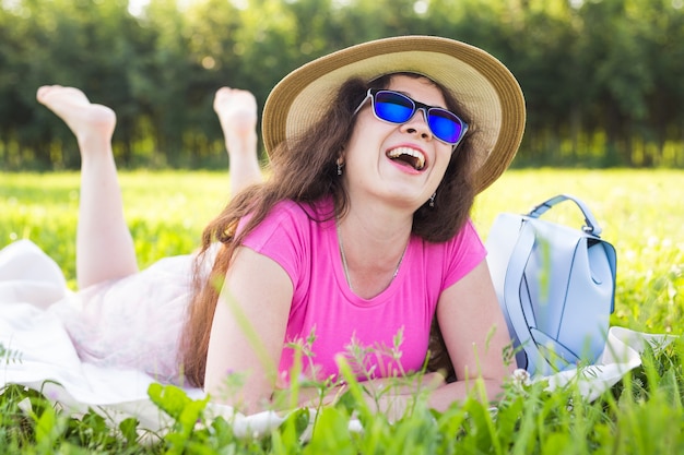 Porträt einer schönen jungen Frau mit Hut und Sonnenbrille, die beim Picknick im Park liegt?