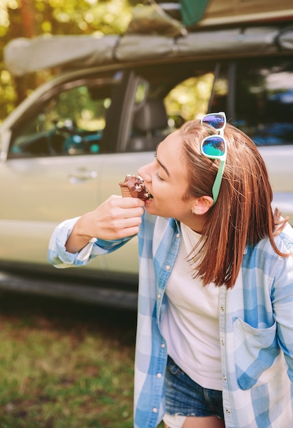 Porträt einer schönen jungen Frau mit kariertem Hemd, die Schokoladeneis gegen das Auto im Wald isst