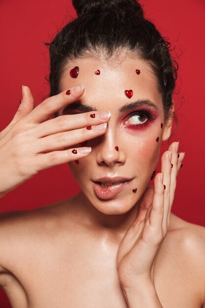 Porträt einer schönen jungen Frau mit rotem hellem Make-up lokalisiert auf roter Wand, die mit Herzen auf Gesicht aufwirft, beißen ihre Lippe.