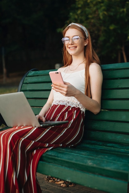 Porträt einer schönen jungen Frau mit roten Haaren und Sommersprossen, die Kamera lachend betrachten, während sie ein Smartphone und einen Laptop auf ihren Beinen im Park halten.