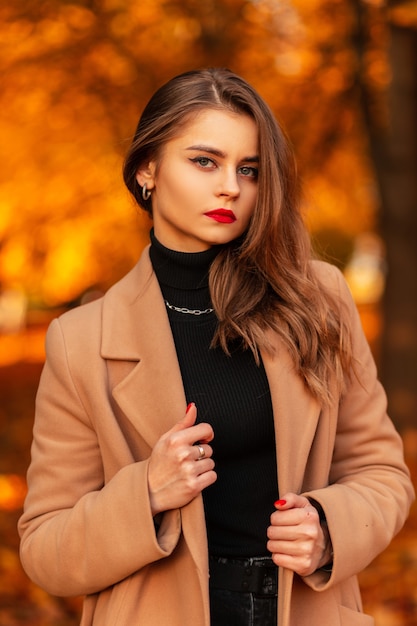 Porträt einer schönen jungen Frau mit roten Lippen in einem modischen beigen Mantel mit einem Pullover in einem Park mit farbigem gelbem Herbstlaub. Weiblicher lässiger Stil und Schönheit