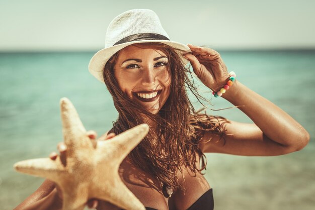 Porträt einer schönen jungen Frau mit Sommerhut, die am Strand genießt. Sie hält Seesterne und schaut mit einem Lächeln auf ihrem Gesicht in die Kamera.