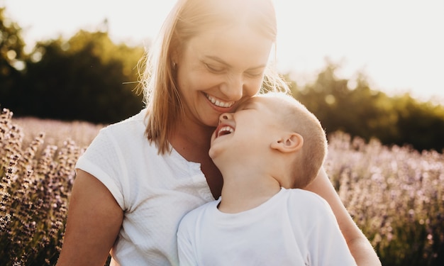 Porträt einer schönen jungen Mutter, die lacht, während sie an ihren Beinen ihr kleines Kind hält, das lacht, während geschlossene Augen in einem Blumenfeld.