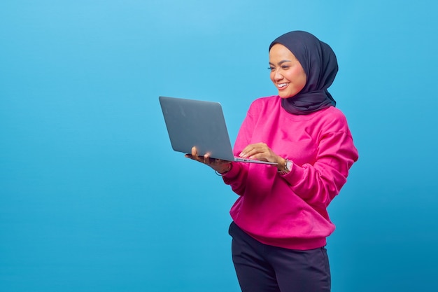 Porträt einer schönen jungen Studentin mit Laptop lächelnd in rosa Jacke auf blauem Hintergrund isoliert