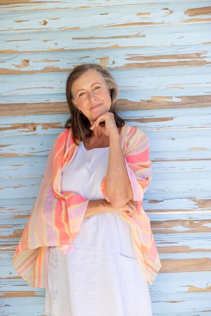 Porträt einer schönen kaukasischen Seniorin mit der Hand am Kinn, die vor einer Holzwand steht. Kopierraum, unverändert, Ruhestand, Urlaub, Schal, Lebensstil, Lächeln und Genusskonzept.