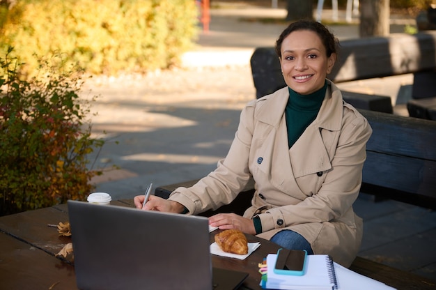 Porträt einer schönen lächelnden afroamerikanischen Frau, die in die Kamera schaut, auf dem Notizblock schreibt, vor einem Laptop-Monitor sitzt und während der Sperrung im Straßencafé auf dem Land aus der Ferne arbeitet