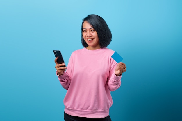 Porträt einer schönen lächelnden asiatischen Frau, die Handy benutzt und Kreditkarte auf blauem Hintergrund hält