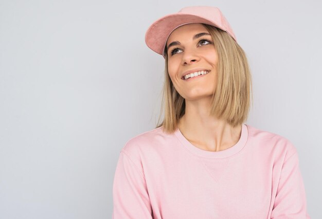 Porträt einer schönen lächelnden blonden Frau mit weißen Zähnen mit attraktivem Aussehen, die rosafarbene Kleidung und eine Kappe trägt, die im Studio gegen eine weiße Wand posiert Glückliche Frau, die positive Emotionen zeigt
