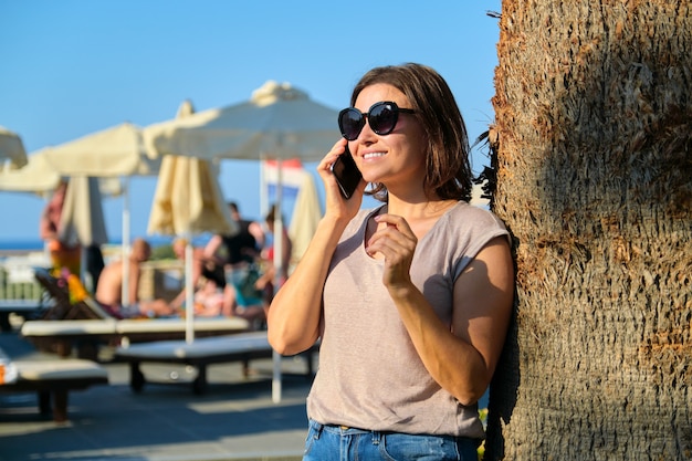 Porträt einer schönen lächelnden reifen Frau, die sich im Resorthotel entspannt, eine Frau mit Sonnenbrille, die in der Nähe der Palme mit Smartphone steht, Kopierraum. Urlaub, Sommer, Tourismus, Resort, Menschen mittleren Alters