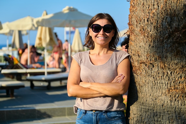 Porträt einer schönen lächelnden reifen Frau, die sich im Resorthotel entspannt, Frau mit Sonnenbrille, die in der Nähe von Palmen mit verschränkten Armen steht, Kopienraum. Urlaub, Sommer, Tourismus, Resort, Menschen mittleren Alters