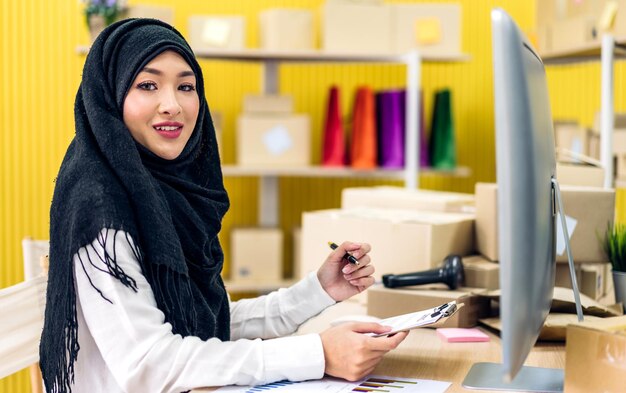 Porträt einer schönen muslimischen Besitzerin, asiatische Frau, Freiberuflerin, KMU-Geschäft, Online-Shopping, Arbeiten und Blick auf die Camara, die zu Hause eine Paketbox auf dem Tisch hält. Geschäftlicher Online-Versand und -Lieferung