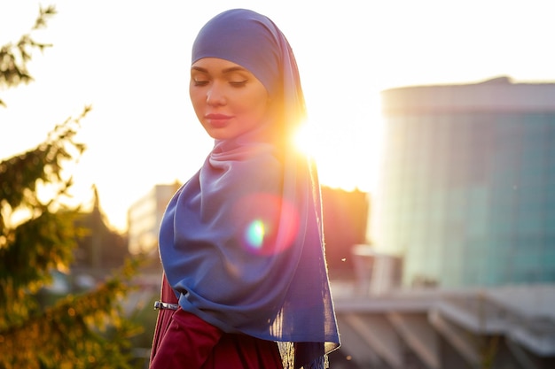 Porträt einer schönen muslimischen Frau mit grünen Augen, die ein blaues Schalgesicht trägt, das mit einem Schleier im Hintergrund bedeckt ist, Waldbäume im Park, Sonnenstrahlen, Sonnenuntergang