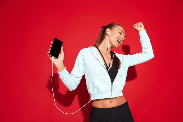 Porträt einer schönen passenden jungen Sportlerin stehend, Musik mit Kopfhörern hörend, Handy haltend