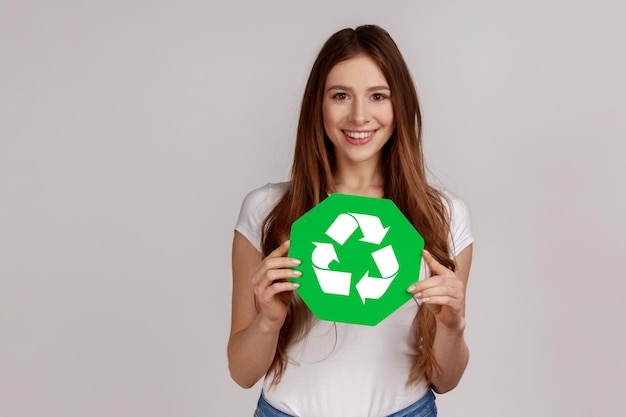 Porträt einer schönen positiven Frau, die grünes Recycling-Symbol hält, Müll sortiert, Umwelt rettet, Ökologie trägt und weißes T-Shirt trägt. Studioaufnahme im Innenbereich isoliert auf grauem Hintergrund.