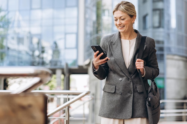 Porträt einer schönen, reifen Geschäftsfrau in Anzug und grauer Jacke, die lächelt und am Telefon auf dem modernen urbanen Hintergrund spricht