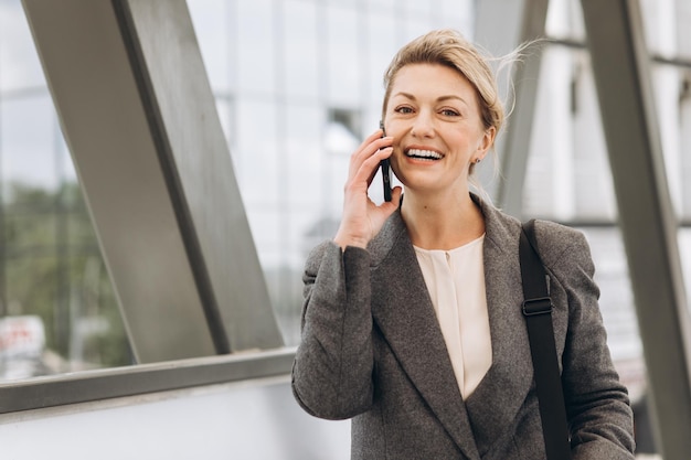 Porträt einer schönen, reifen Geschäftsfrau in Anzug und grauer Jacke, die lächelt und am Telefon auf dem modernen urbanen Hintergrund spricht