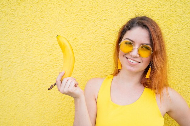 Porträt einer schönen rothaarigen Frau mit Sonnenbrille auf gelbem Hintergrund Attraktives Mädchen posiert mit einer Banane in den Händen