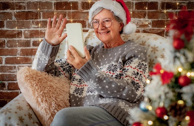 Porträt einer schönen Seniorin mit Weihnachtsmütze, die zur Weihnachtszeit per Handy chattet. Ältere lächelnde Dame, die allein zu Hause mit drahtloser Technologie sitzt