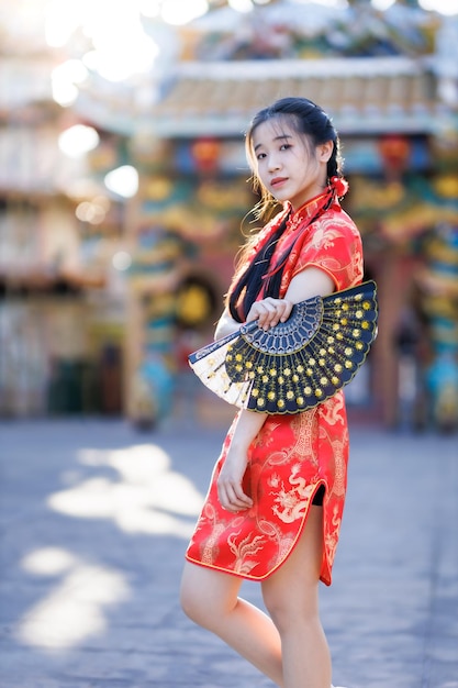 Porträt einer schönen, süßen lächelnden asiatischen jungen Frau, die rote traditionelle chinesische Cheongsam-Dekoration trägt und einen chinesischen Fanning für das chinesische Neujahrsfest am chinesischen Schrein in Thailand hält