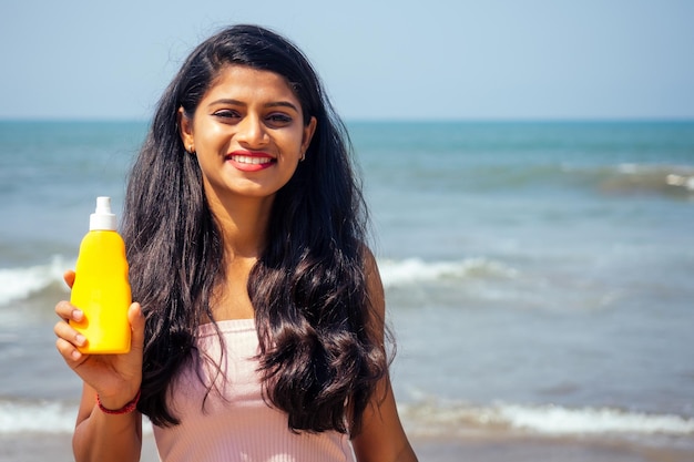 Porträt einer schönen und lächelnden schneeweißen lächelnden inderin mit schwarzen haaren und dunkler haut in einem rosa t-shirt, das eine flasche sonnenschutzspray auf beachgirl hält, das spf-körperparadiesurlaub genießt