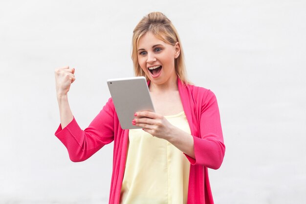 Porträt einer schönen, unglaublichen jungen Frau in rosafarbener Bluse, die ihr Tablet mit überraschtem Gesicht und erhobener Hand steht und beobachtet und benutzt. Innen, isoliert, Studioaufnahme, weißer Hintergrund