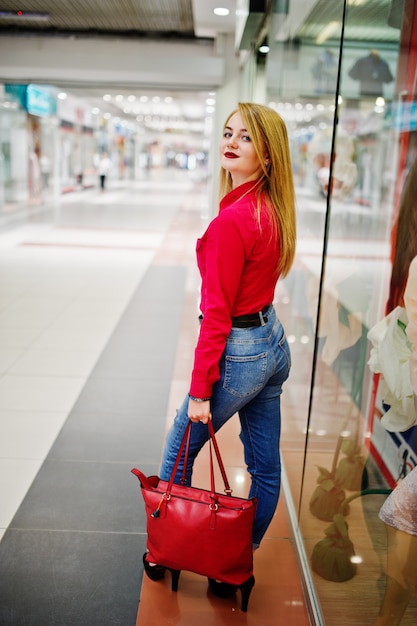 Porträt einer Schönheit, die rote Bluse, zufällige Jeans und schwarze hohe Absätze trägt, wirft mit roter Lederhandtasche im enormen Einkaufszentrum auf.